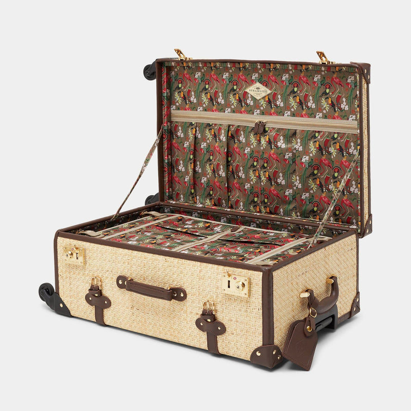 Open product view of the check-in spinner Explorer rattan suitcase in natural wicker body and brown trims with exotic bird print lining and brown leather luggage tag